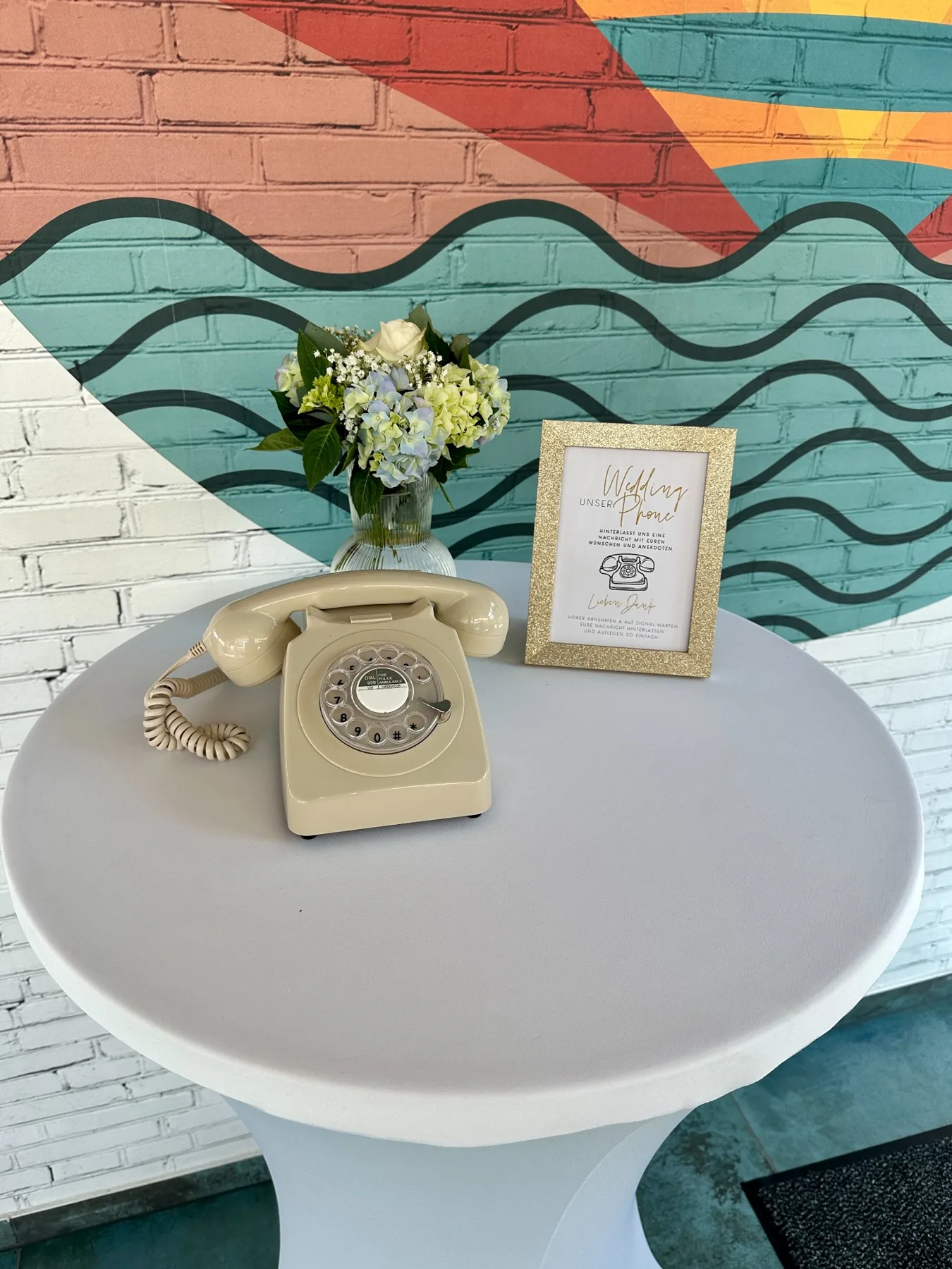 Cremefarbenes Weddingphone auf einem runden Tisch mit Blumen und Hinweisschild